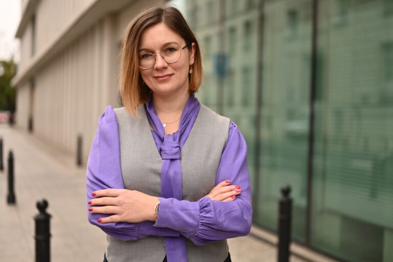 Portret posłanki Pauliny Matysiak, stoi ze skrzyżowanymi rękoma, w szarej kamizelce i fioletowej koszuli. W tle budynek U przy ul. Wiejskiej.