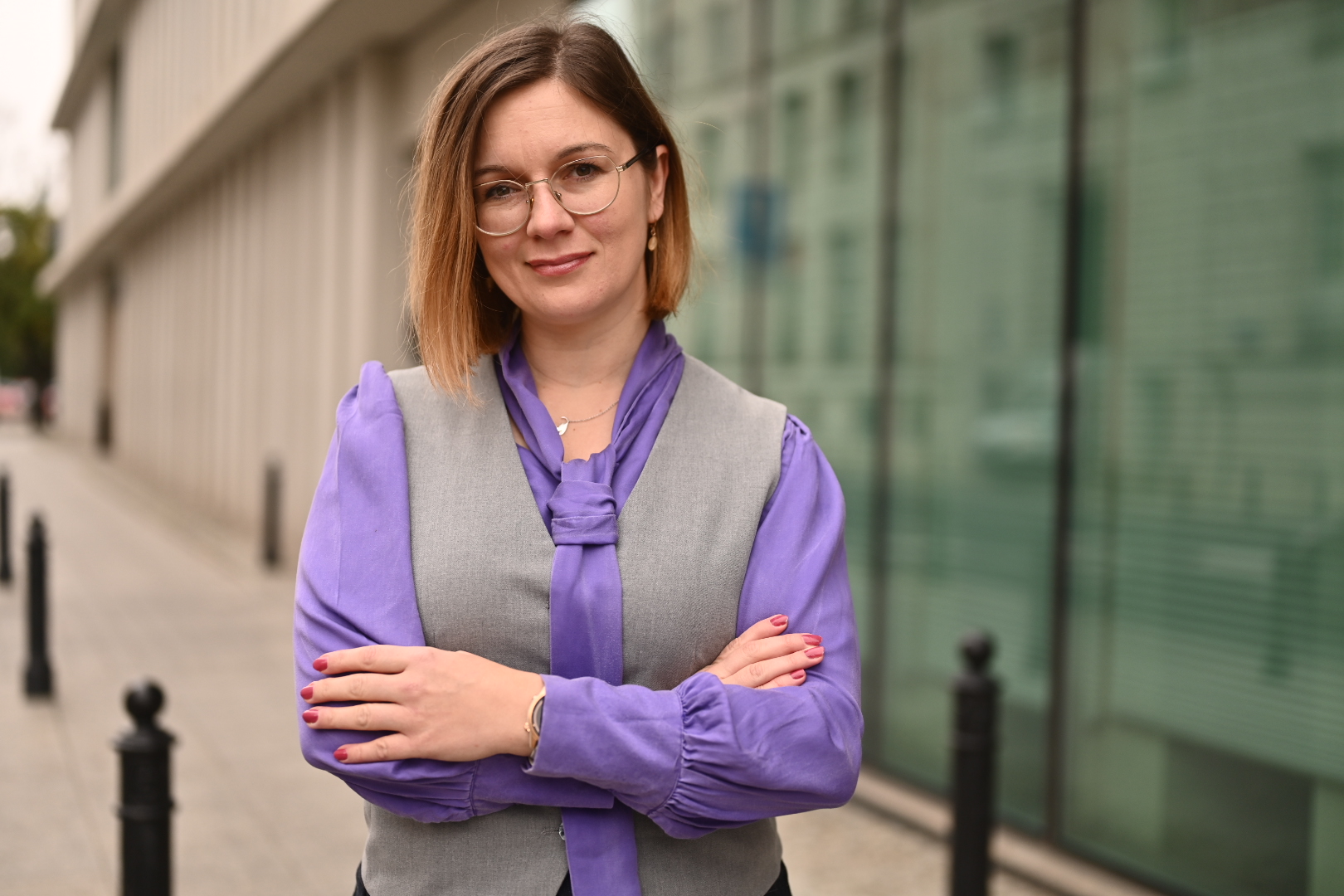 Portret posłanki Pauliny Matysiak, stoi ze skrzyżowanymi rękoma, w szarej kamizelce i fioletowej koszuli. W tle budynek U przy ul. Wiejskiej.