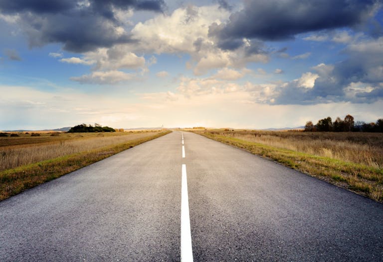 A long straight road stretches through fields under a dramatic cloudy sky.