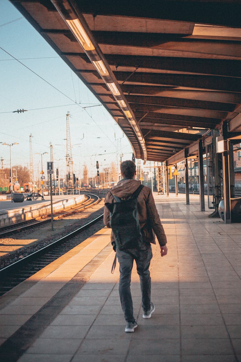 A man with a backpack walks on an empty railway platform. Sunlight casts warm tones across the scene.