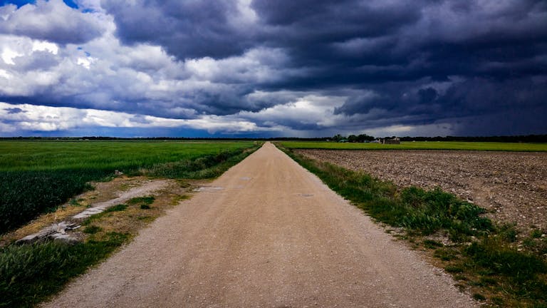 A dirt road stretches through lush green fields under a dramatic cloudy sky.