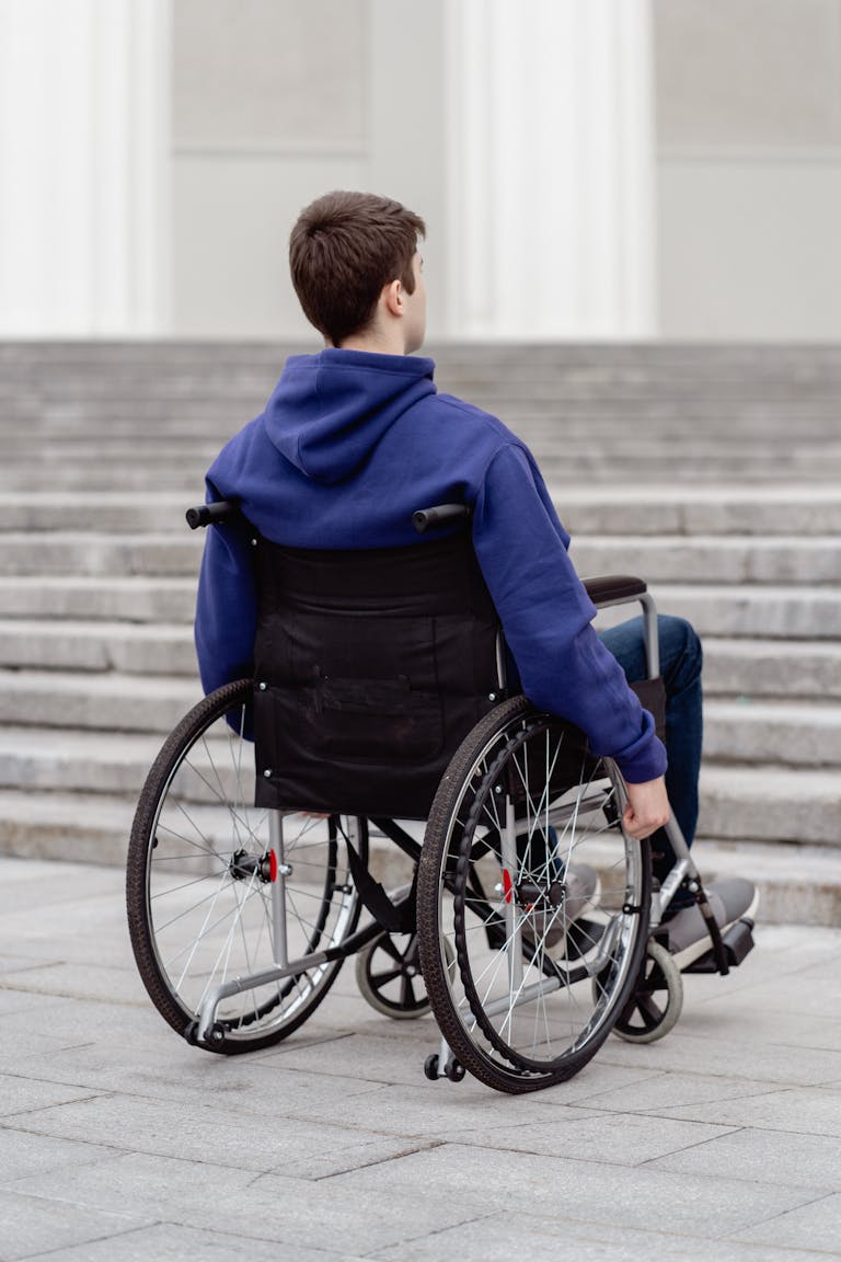 A young man in a wheelchair with a blue hoodie facing a staircase outdoors.