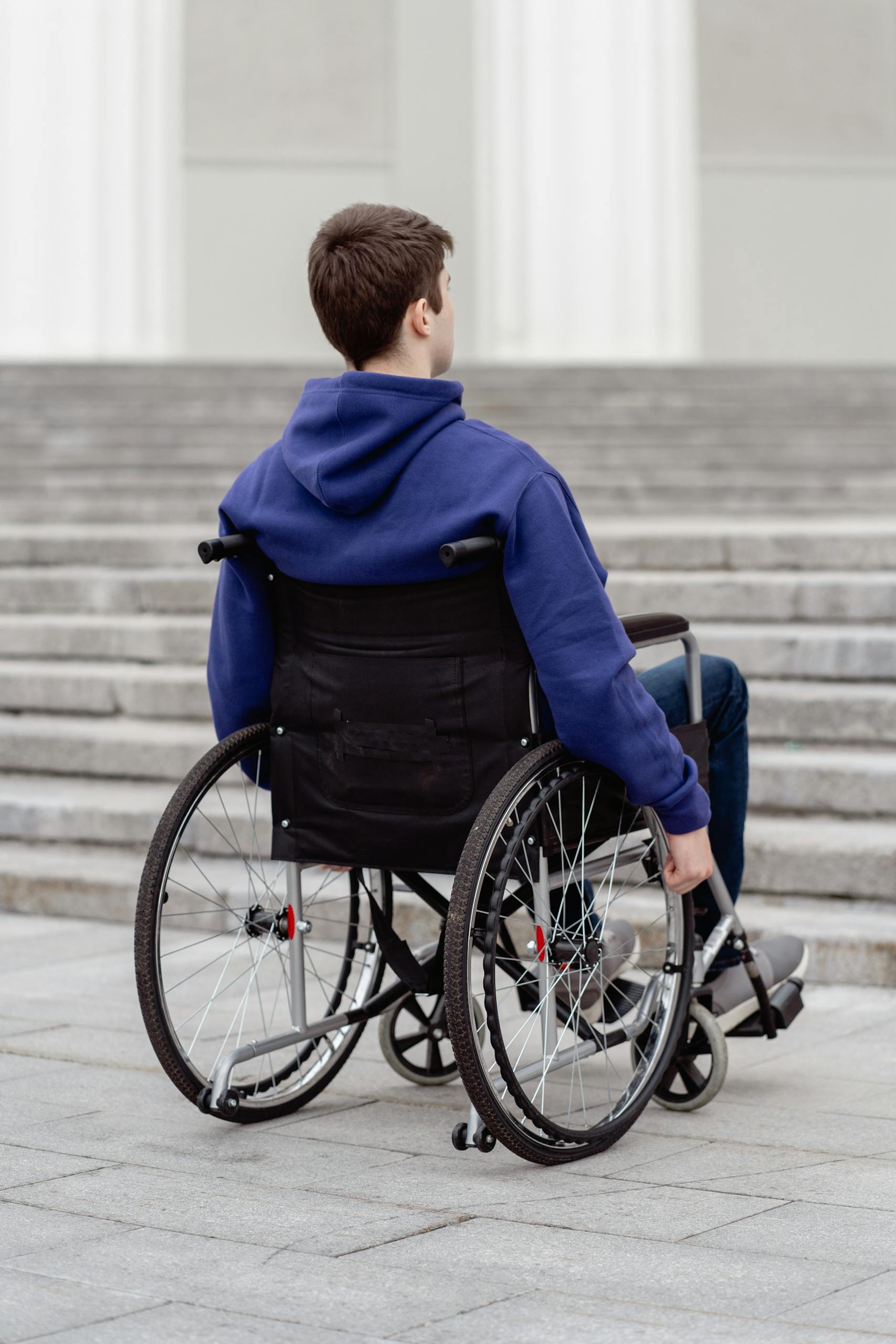 A young man in a wheelchair with a blue hoodie facing a staircase outdoors.