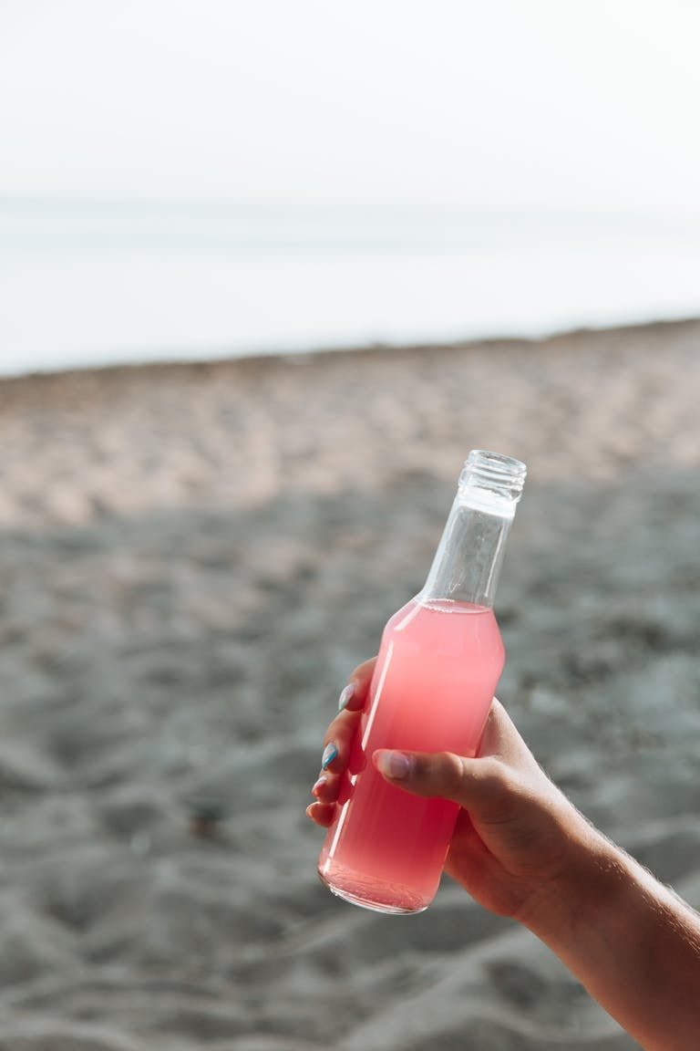 Close-up of a hand holding a pink drink in a glass bottle on a sandy beach.