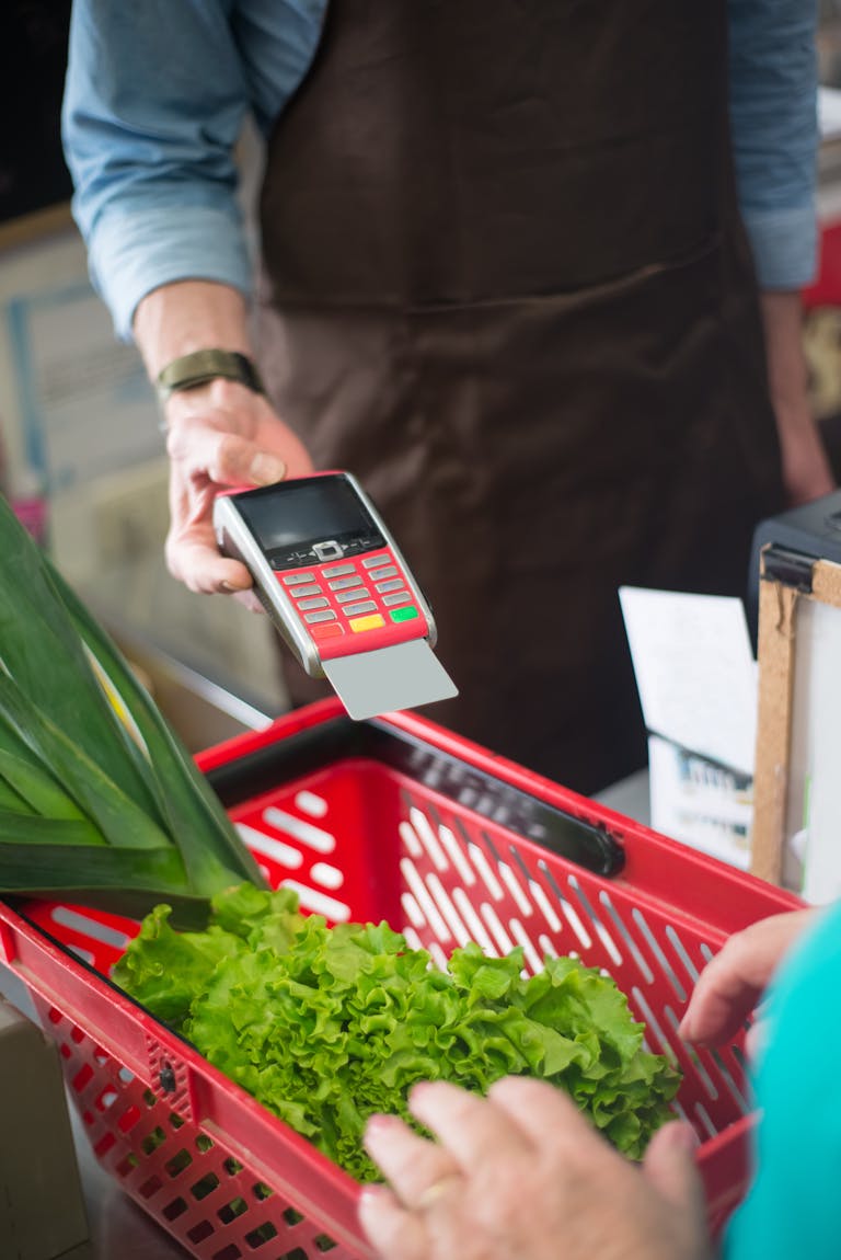 A customer completes a contactless payment at a grocery store checkout, featuring fresh produce in a red basket.