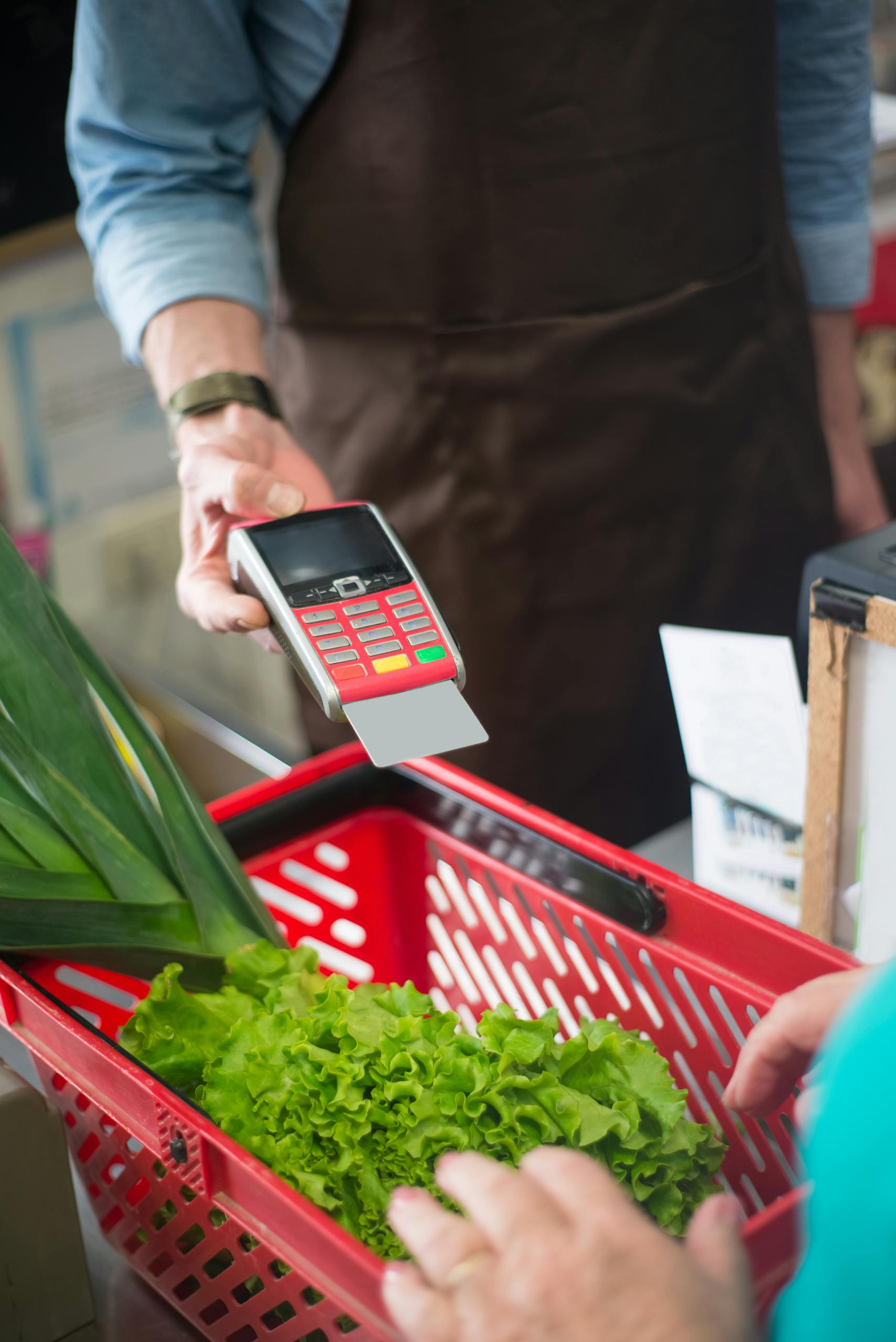 A customer completes a contactless payment at a grocery store checkout, featuring fresh produce in a red basket.