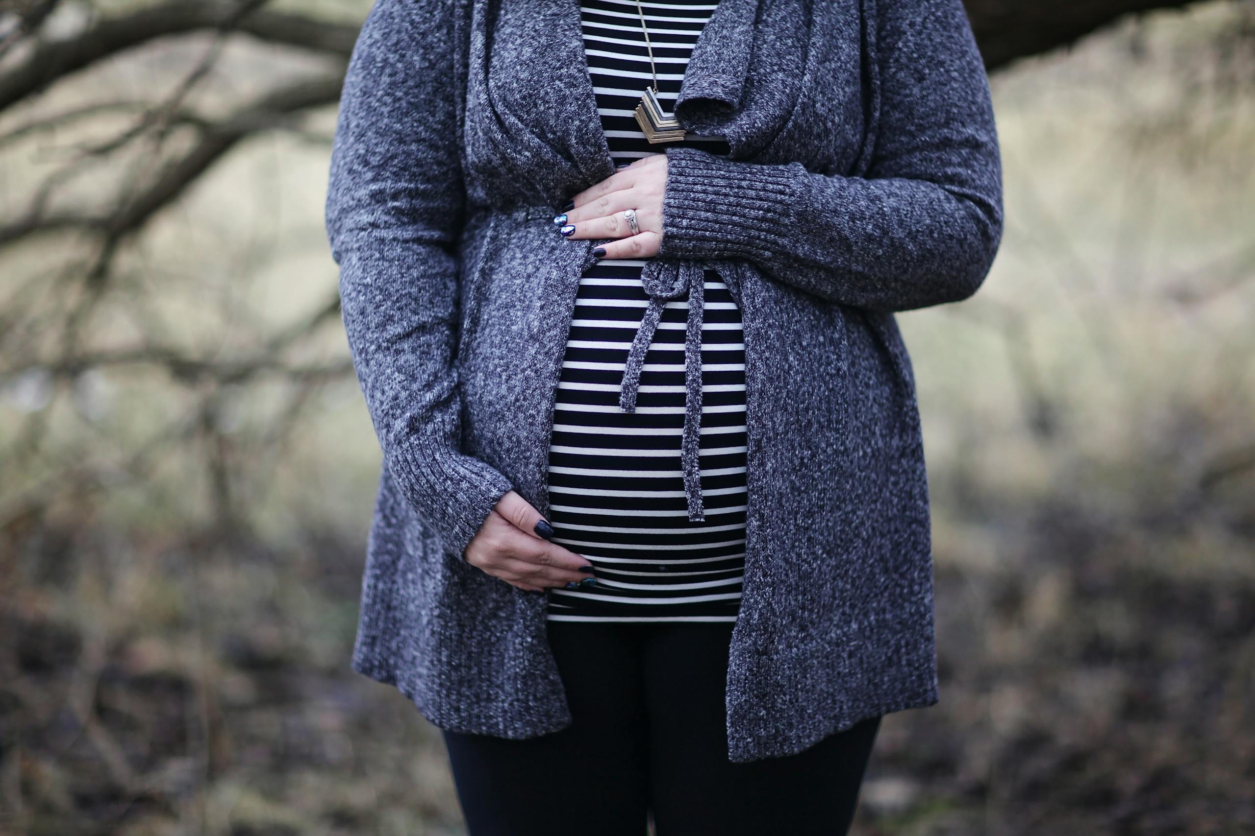 Pregnant woman in striped shirt and cozy sweater outdoors, embracing motherhood.
